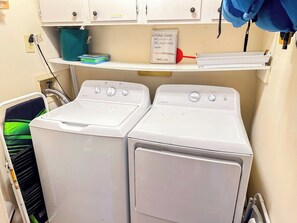 A laundry area with two front-loading washing machines, a shelf with a sign, and a green bucket. - Tilghman Beach and Racquet Club 323 (North Myrtle Beach)