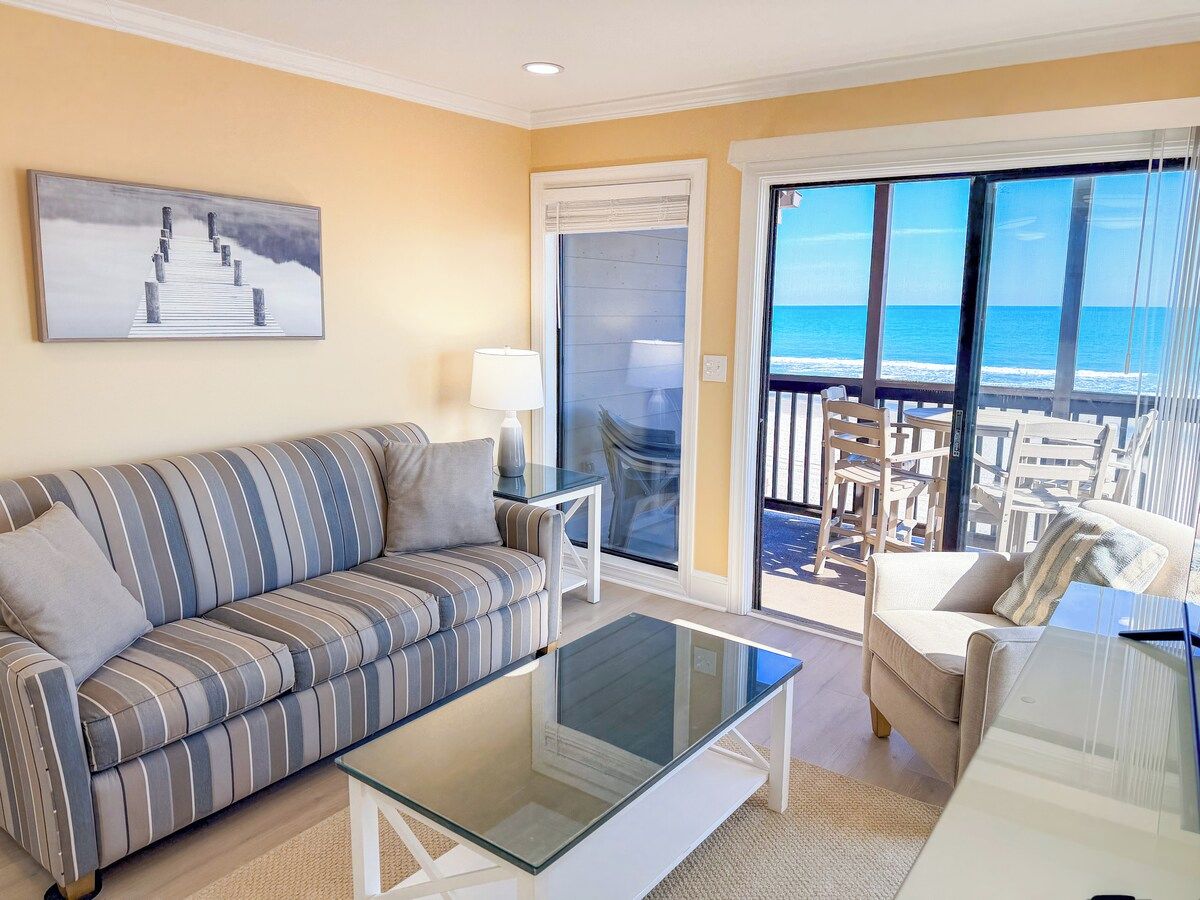 A living room with a sofa, a coffee table, and a view of the ocean through large windows.