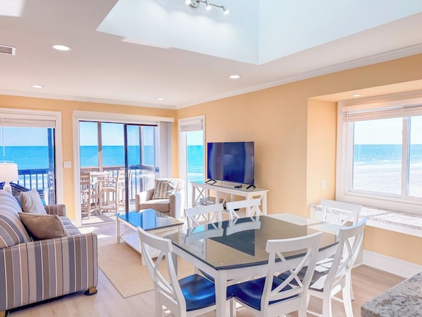 A modern living room with a glass dining table, white chairs, a sofa, and a flat-screen TV.