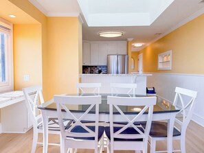 A dining area with a glass table and white chairs, a kitchen in the background, and a window with blinds.
