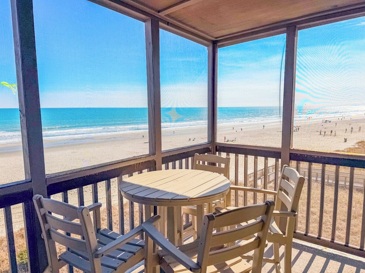 A covered outdoor seating area with a table and chairs, overlooking a beach with people and a clear sky.