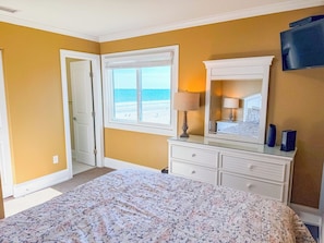 Een slaapkamer met een bed, een dressoir, een televisie en een raam met uitzicht op het strand.