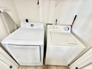 Uma área de lavanderia com duas máquinas de lavar brancas e uma secadora, uma toalha branca pendurada em um gancho e uma tomada na parede.
