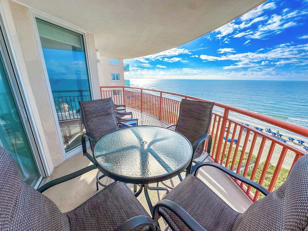 A balcony with a glass table and chairs, overlooking a beach with a red railing.