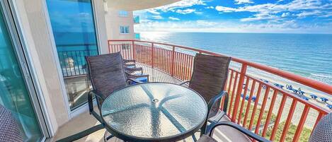 A balcony with a glass table and chairs, overlooking a beach with a red railing.