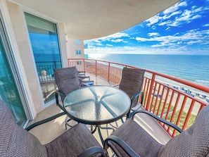 Un balcón con mesa y sillas de vidrio, con vista a una playa con barandilla roja.