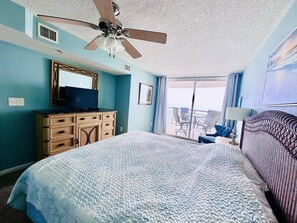 A hotel room with a bed, a ceiling fan, a dresser, a television, and a balcony with chairs.