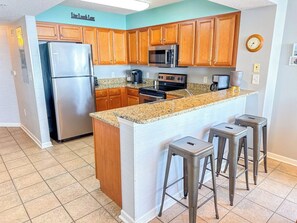 A kitchen with wooden cabinets, a stainless steel refrigerator, a microwave, an oven, and a granite countertop. There are three bar stools and a clock on the wall. - South Shore Villas 801 (North Myrtle Beach)