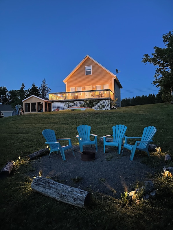 Uma casa com um deck e quatro cadeiras Adirondack azuis dispostas ao redor de um braseiro.
