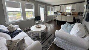A modern living room with a white sofa, a coffee table, and a ceiling fan. There are large windows providing a view of the outdoors. - River Front Property With Water View, Minutes To Brackley, & Screened In Dining (Oyster Bed)