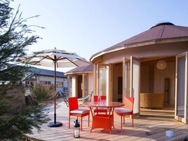 Terraza de madera con una mesa, sillas, una hamaca y un ventanal de vidrio que da a un espacio interior.