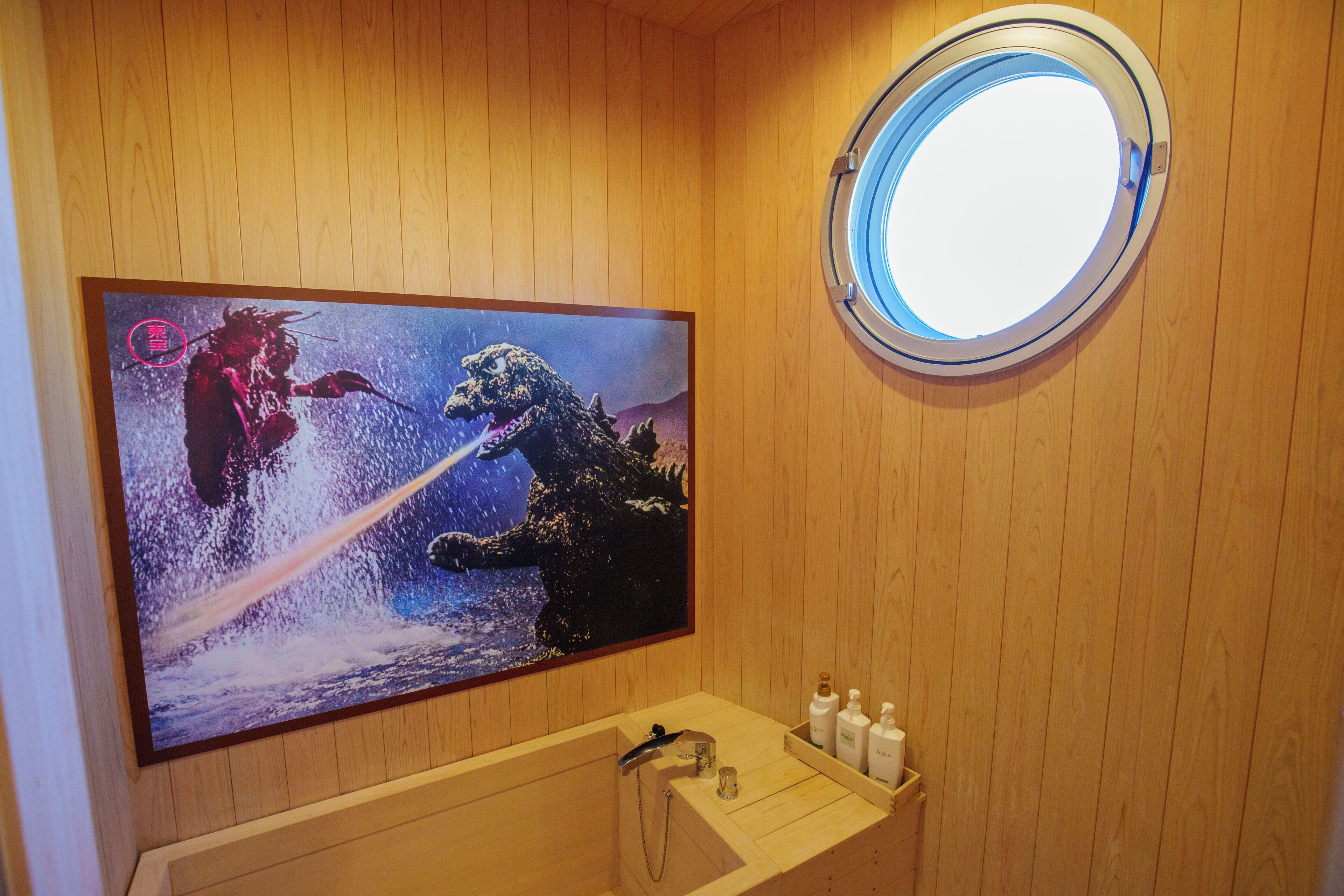 A wooden-paneled room with a large poster of Godzilla and another creature, a round window, and a wooden sink with toiletries.