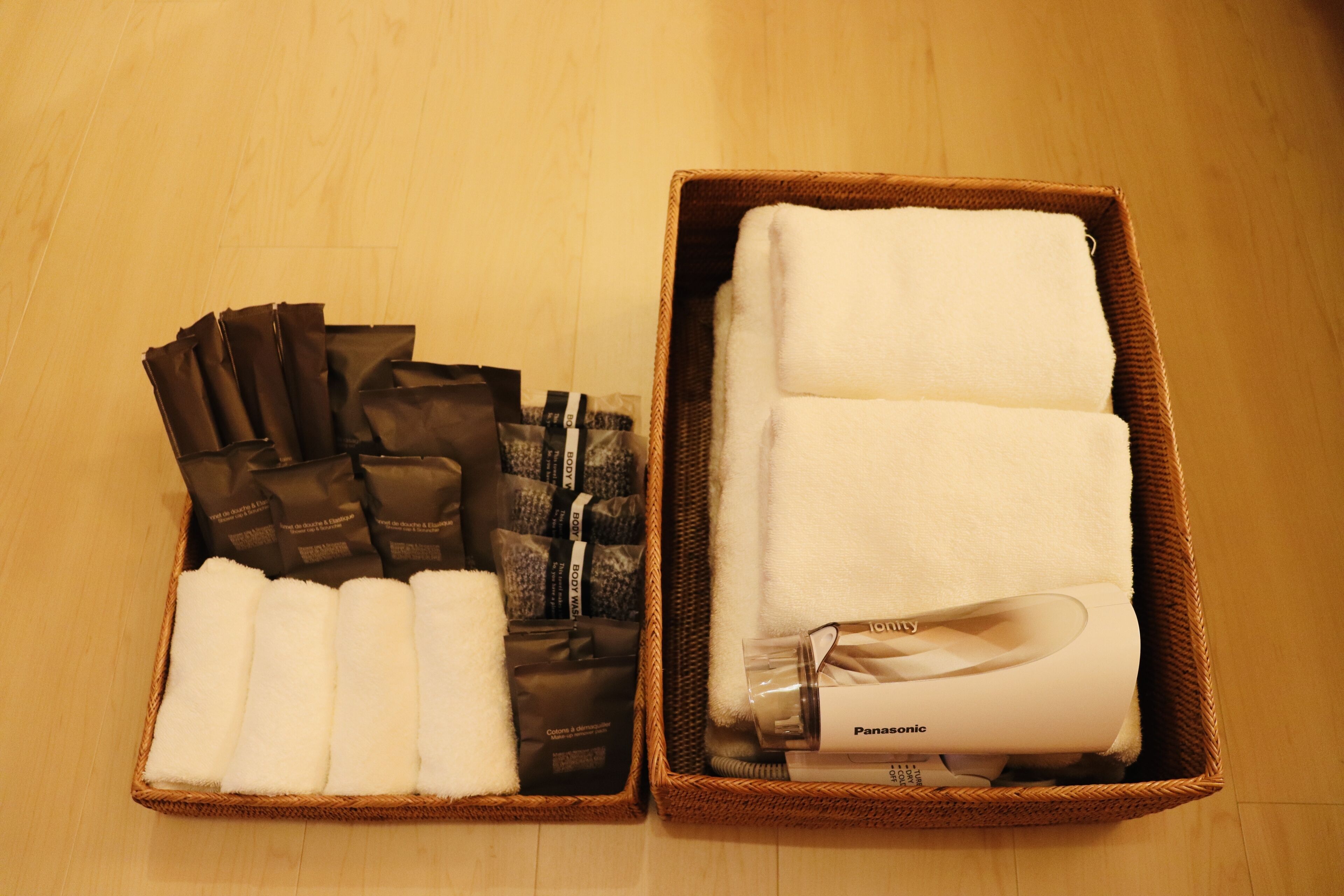 Two wicker baskets on a wooden floor, one containing neatly folded white towels and a bottle labeled "Panasonic," and the other holding several brown bags and folded white towels.