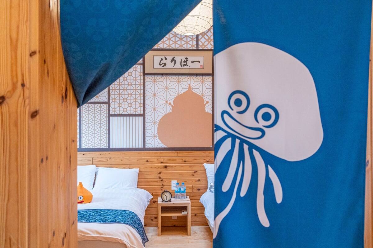 Une chambre avec deux lits, une tête de lit en bois, une petite table de chevet avec une horloge et des bouteilles d’eau, et un mur orné d’un motif géométrique et d’une grande illustration représentant un poulpe.