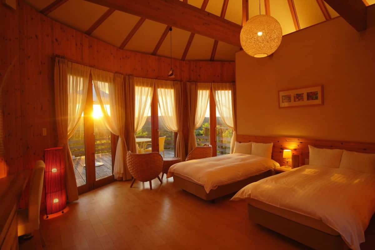 Une chambre confortable avec deux lits, des poutres en bois, de grandes fenêtres avec des rideaux, un fauteuil en osier et un luminaire suspendu.