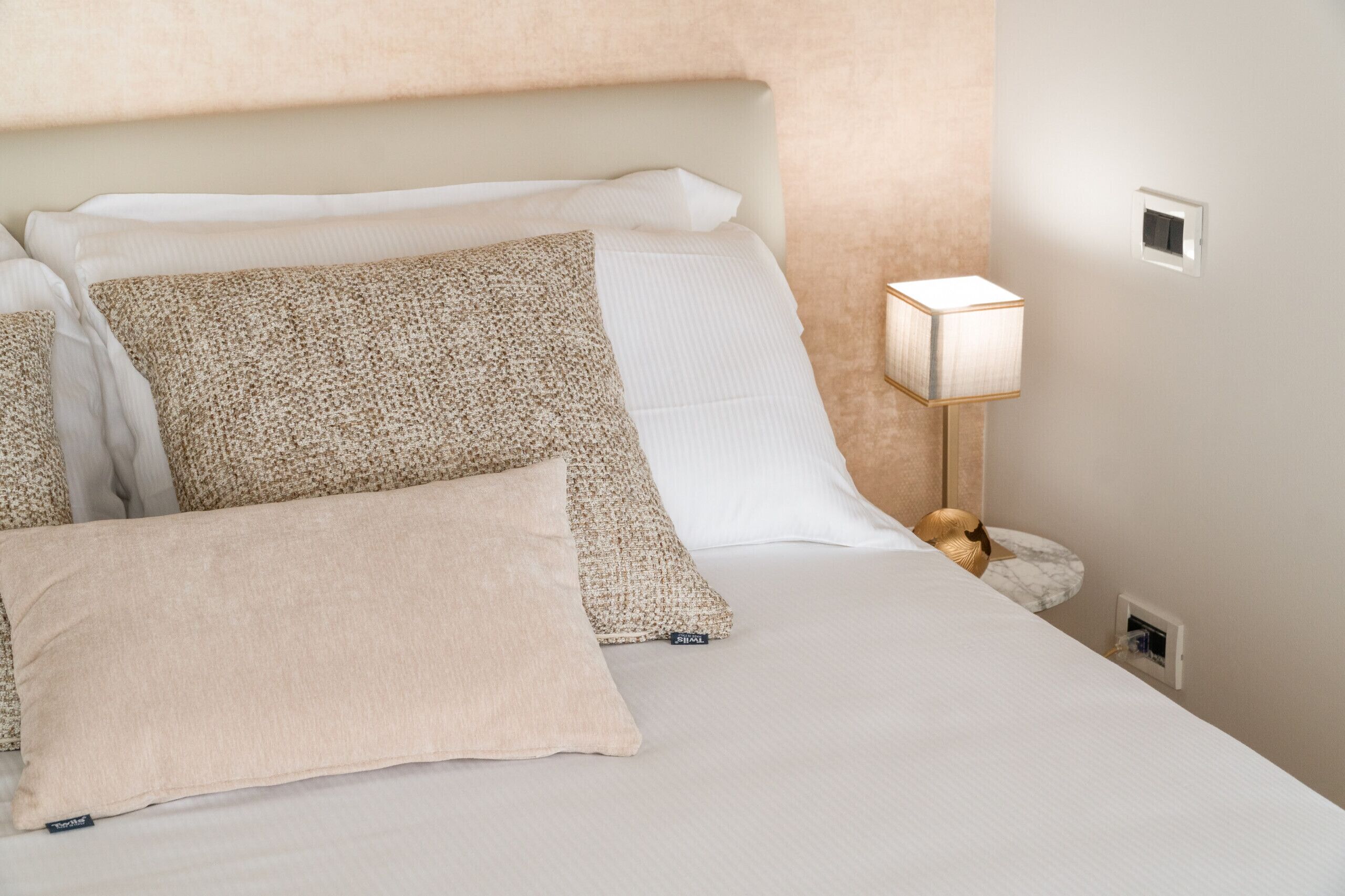 Ein ordentlich bezogenes Bett mit weißer Decke und mehreren Kissen, einer Nachttischlampe und einer Steckdose.