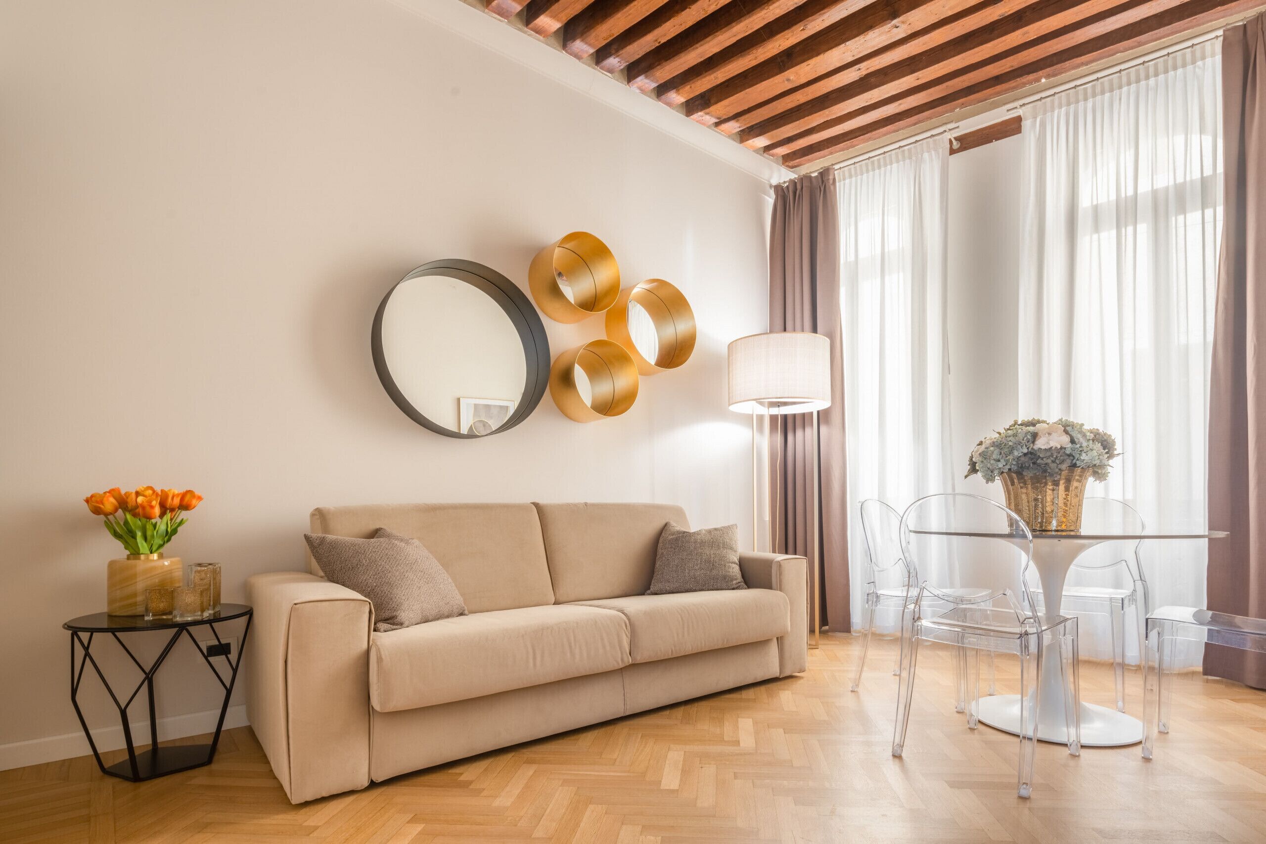 A living room with a beige sofa, a round mirror, and a table with clear chairs.
