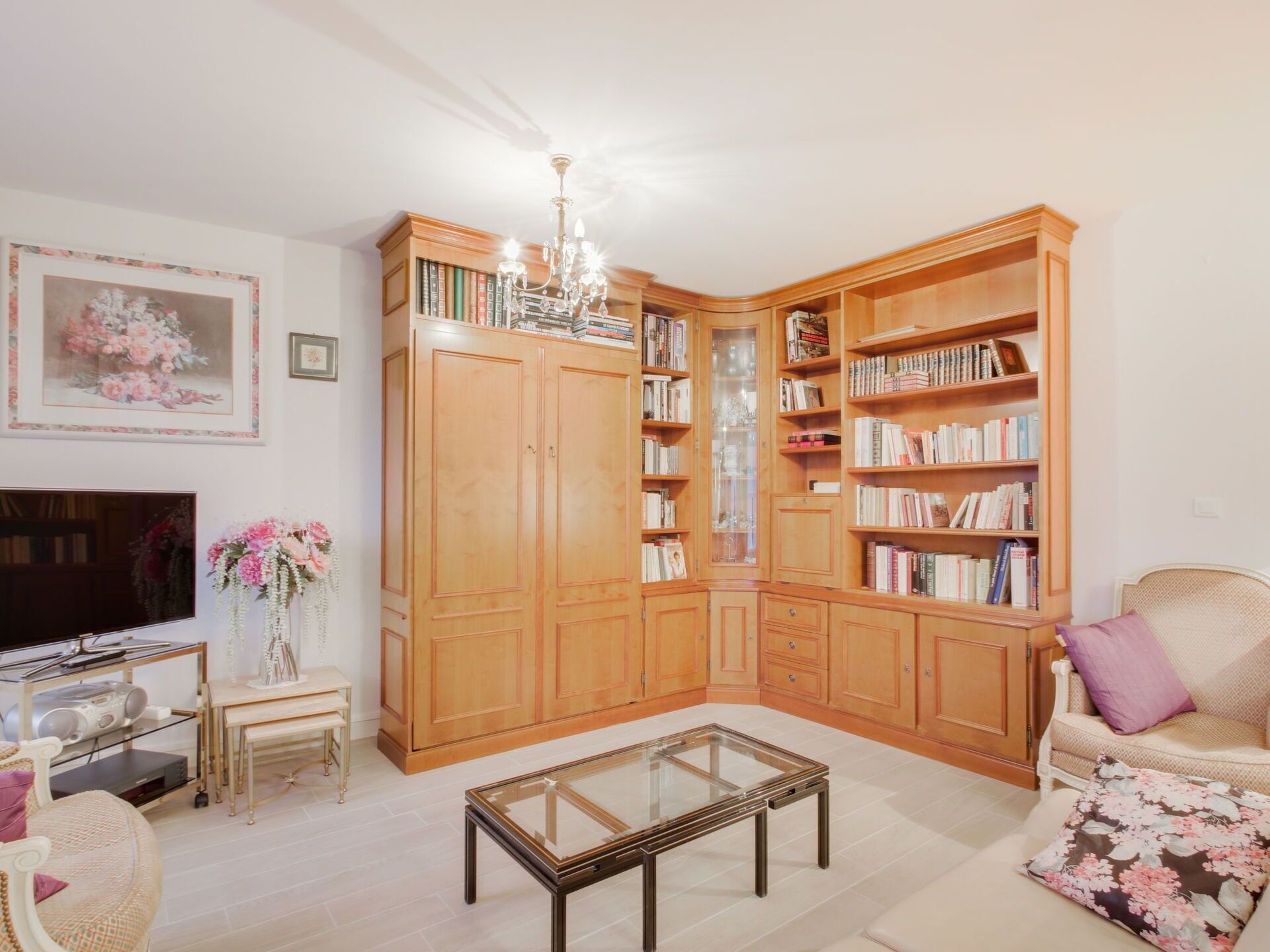 Ein Wohnzimmer mit Fernseher, einem Glastisch, einer Couch, einem Bücherregal und einer Chandelieber.
