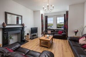 Ein Wohnzimmer mit Kamin, Fernseher, Ledersofas, einem Couchtisch und einem großen Fenster mit Vorhängen.
