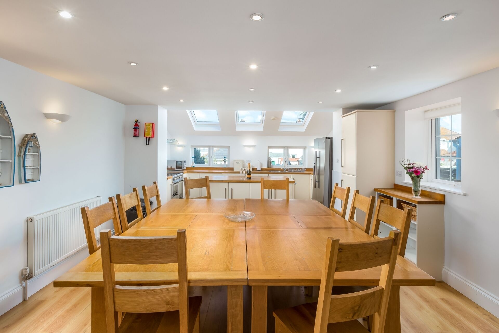 A spacious dining area with a long wooden table and chairs, a kitchen in the background, and skylights on the ceiling.