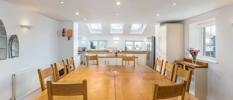A spacious dining area with a long wooden table and chairs, a kitchen in the background, and skylights on the ceiling.