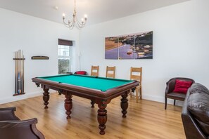 A pool table with a green felt top, wooden legs, and a chandelier above.