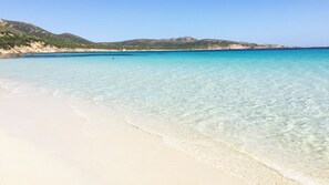 Ein Strand mit klarem türkisblauem Wasser, weißem Sand und Bergen in der Ferne.