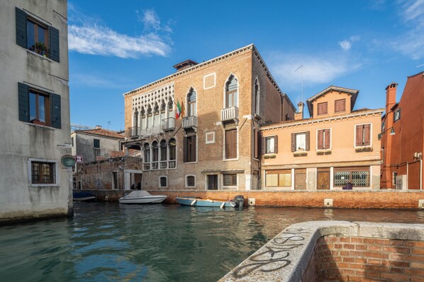 A canal with buildings on either side, including a brick building with arched windows and balconies, and a boat docked nearby.