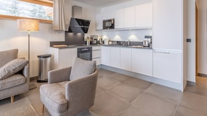 A modern kitchen with white cabinetry, a built-in oven, and a microwave. There is a dining area with a grey sofa and a lamp.