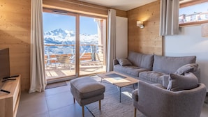 A modern living room with a sofa, ottoman, and coffee table, a TV, and a view of snow-capped mountains through large windows.