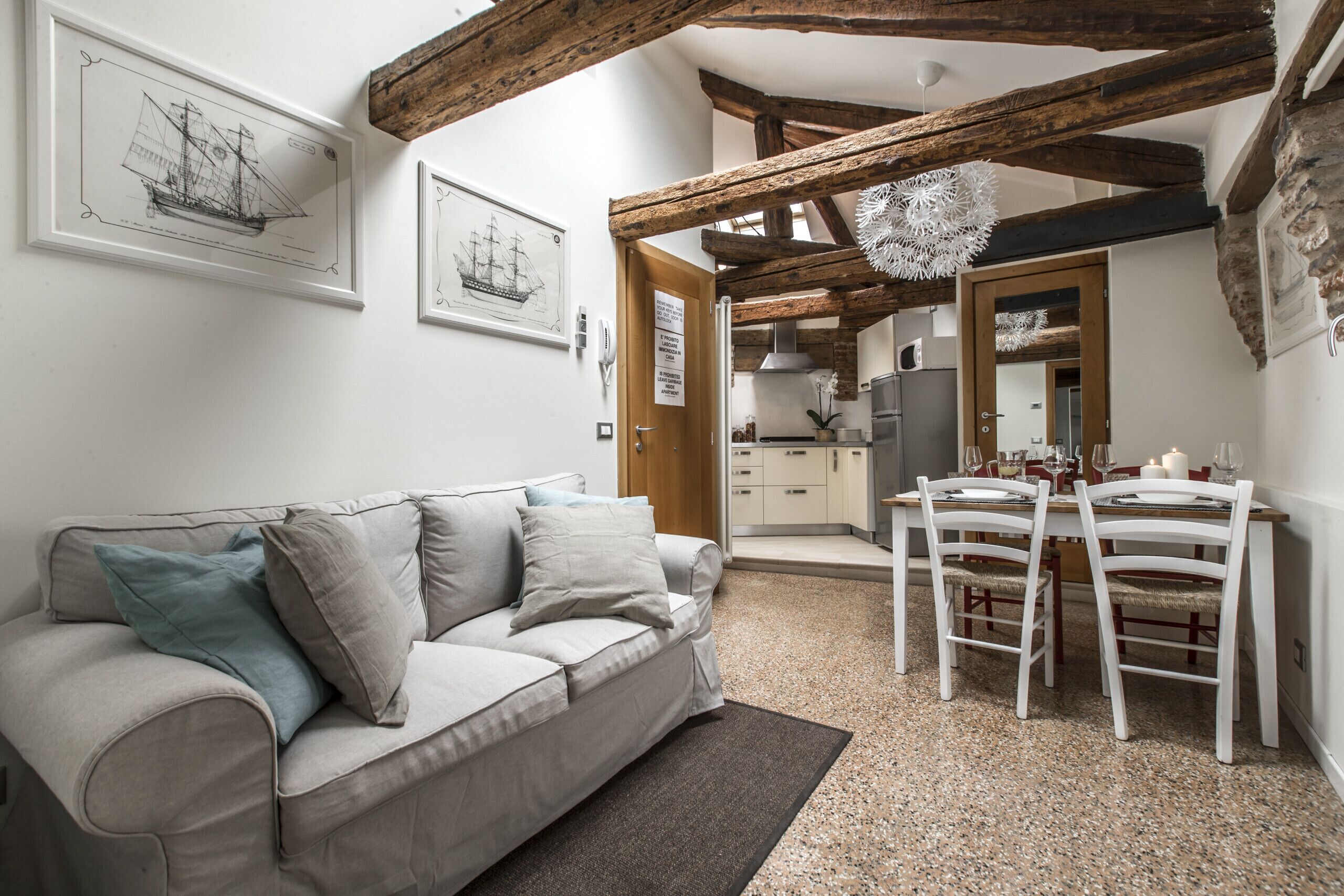 Un salon chaleureux avec un canapé, une table à manger et des chaises. Les murs sont ornés de tableaux encadrés, le plafond est en bois et, au fond, on aperçoit une cuisine.