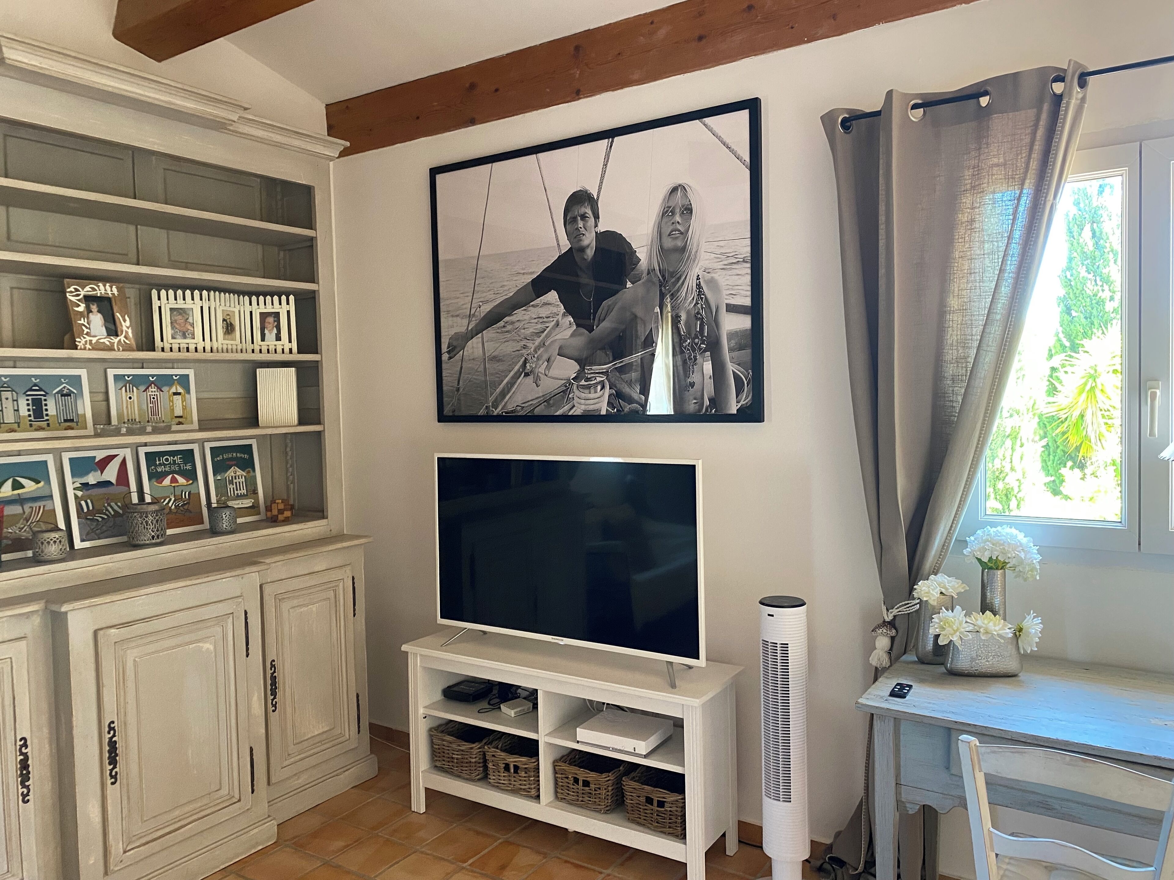 Een woonkamer met een televisie, witte kasten, een lijst met een afbeelding en een raam met gordijnen.