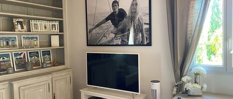 Een woonkamer met een televisie, witte kasten, een lijst met een afbeelding en een raam met gordijnen.