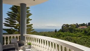 Un balcon agrémenté de meubles en osier, d’une plante en pot et offrant une vue sur la mer et la verdure.