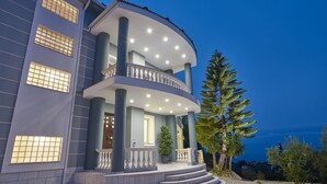Un immeuble à plusieurs étages doté d’un balcon incurvé, d’un revêtement de sol en pierre et d’un grand arbre au premier plan.