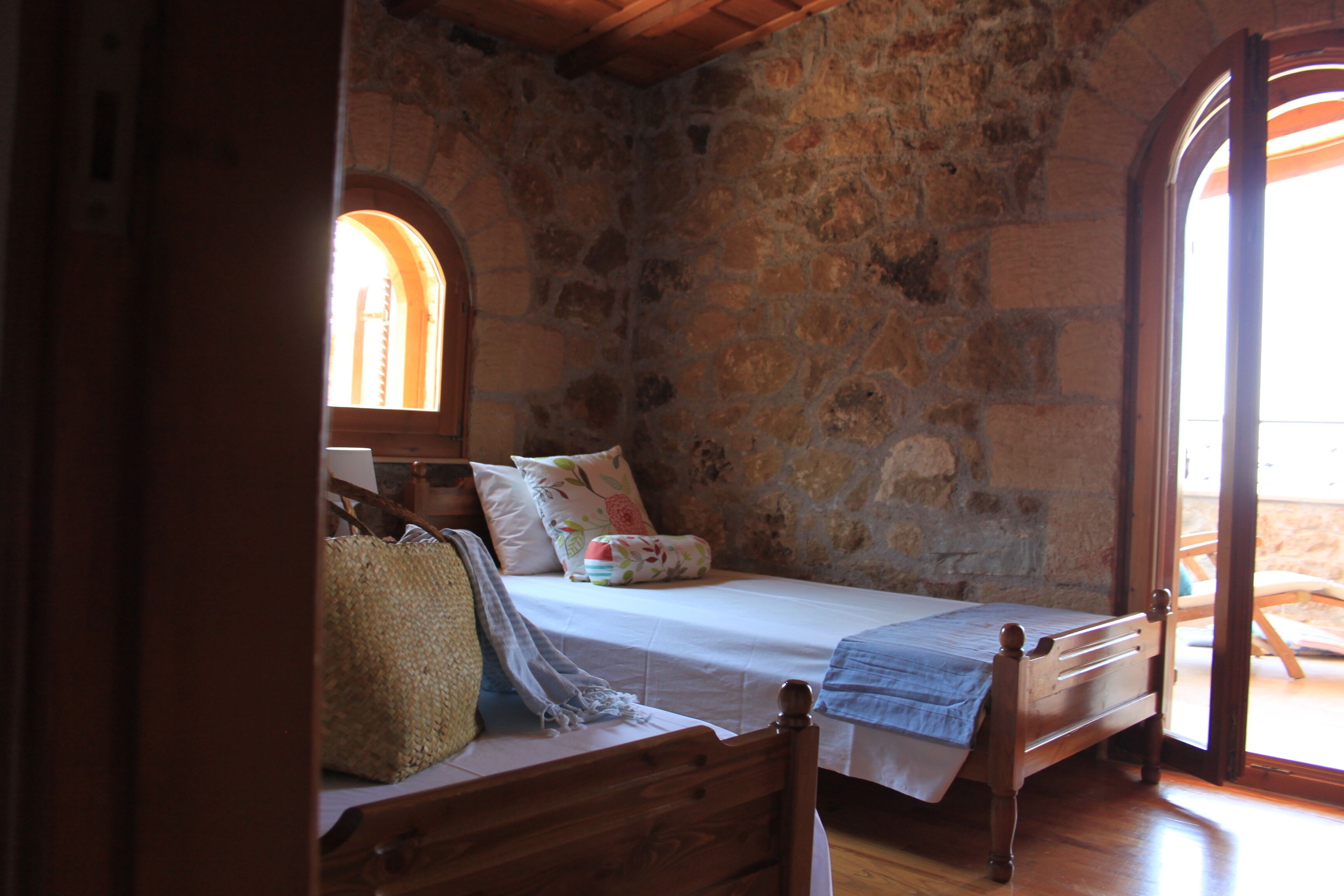 A room with a stone wall, a bed with a wooden headboard, a window, and a door leading outside.