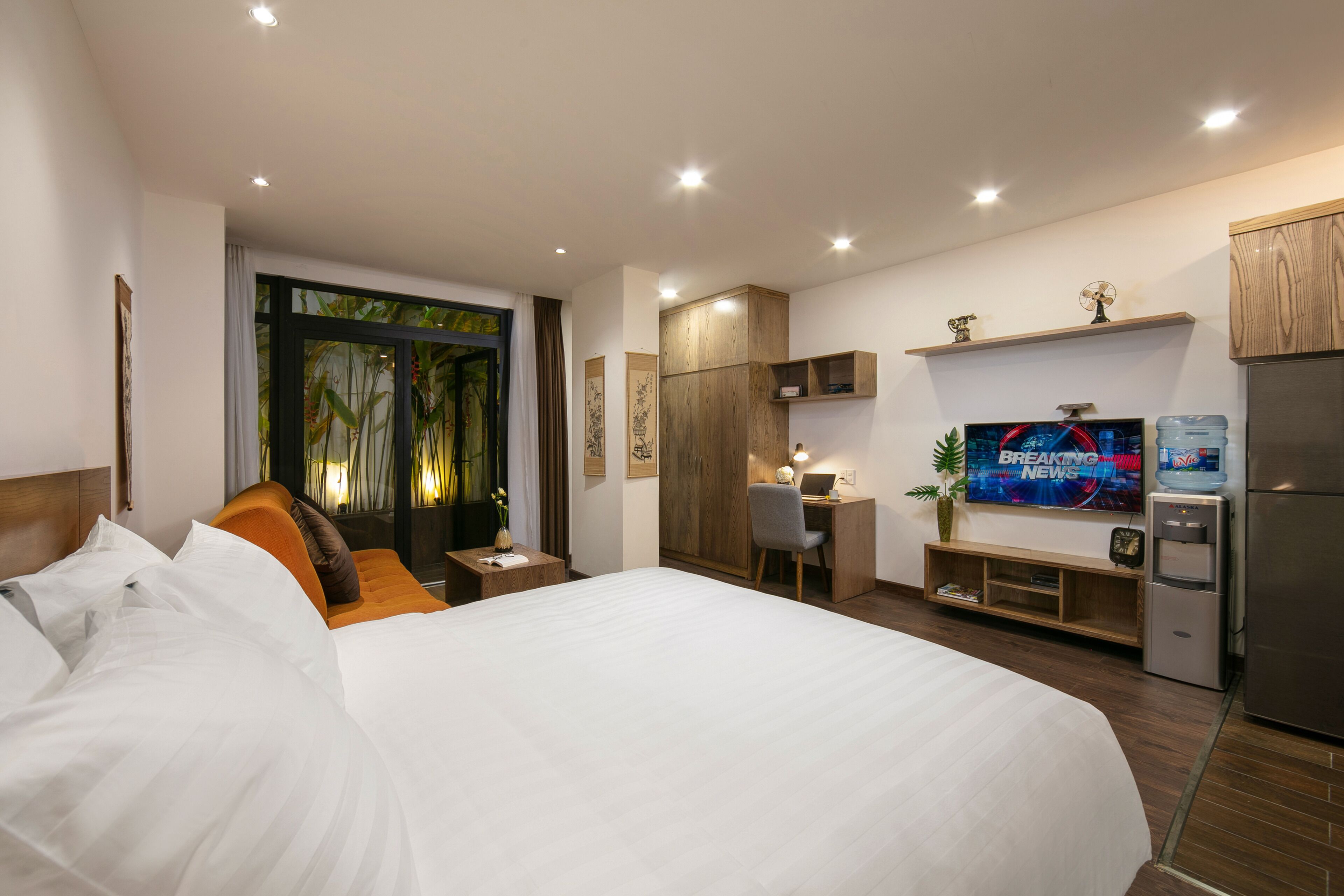 Quarto de hotel moderno com uma cama grande, uma mesa de trabalho com cadeira, televisão e frigorífico.