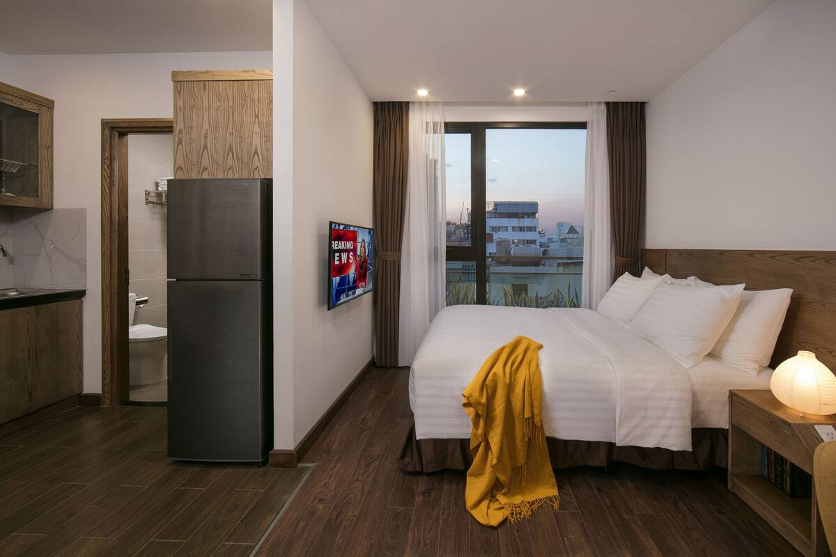 A hotel room with a bed, a bedside table with a lamp, a refrigerator, a wooden cabinet, and a bathroom visible through an open door.