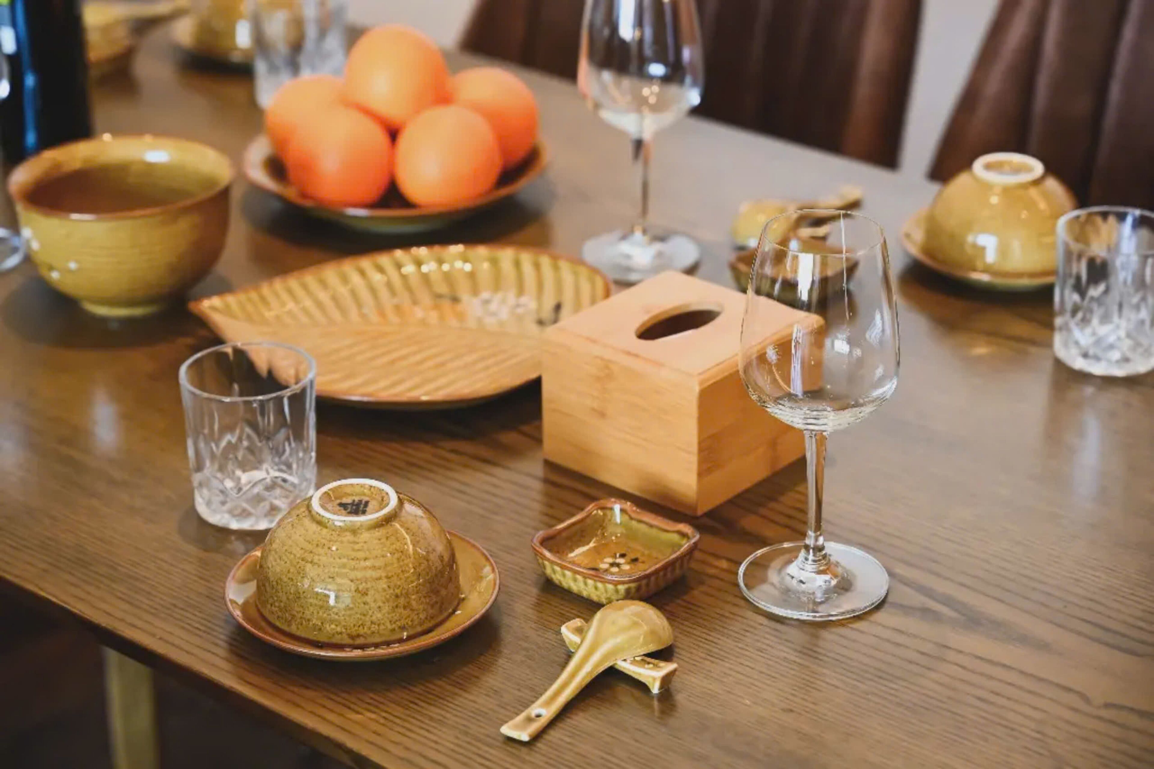 A wooden dining table with a bowl of oranges, a glass, a teapot, and a wooden box.