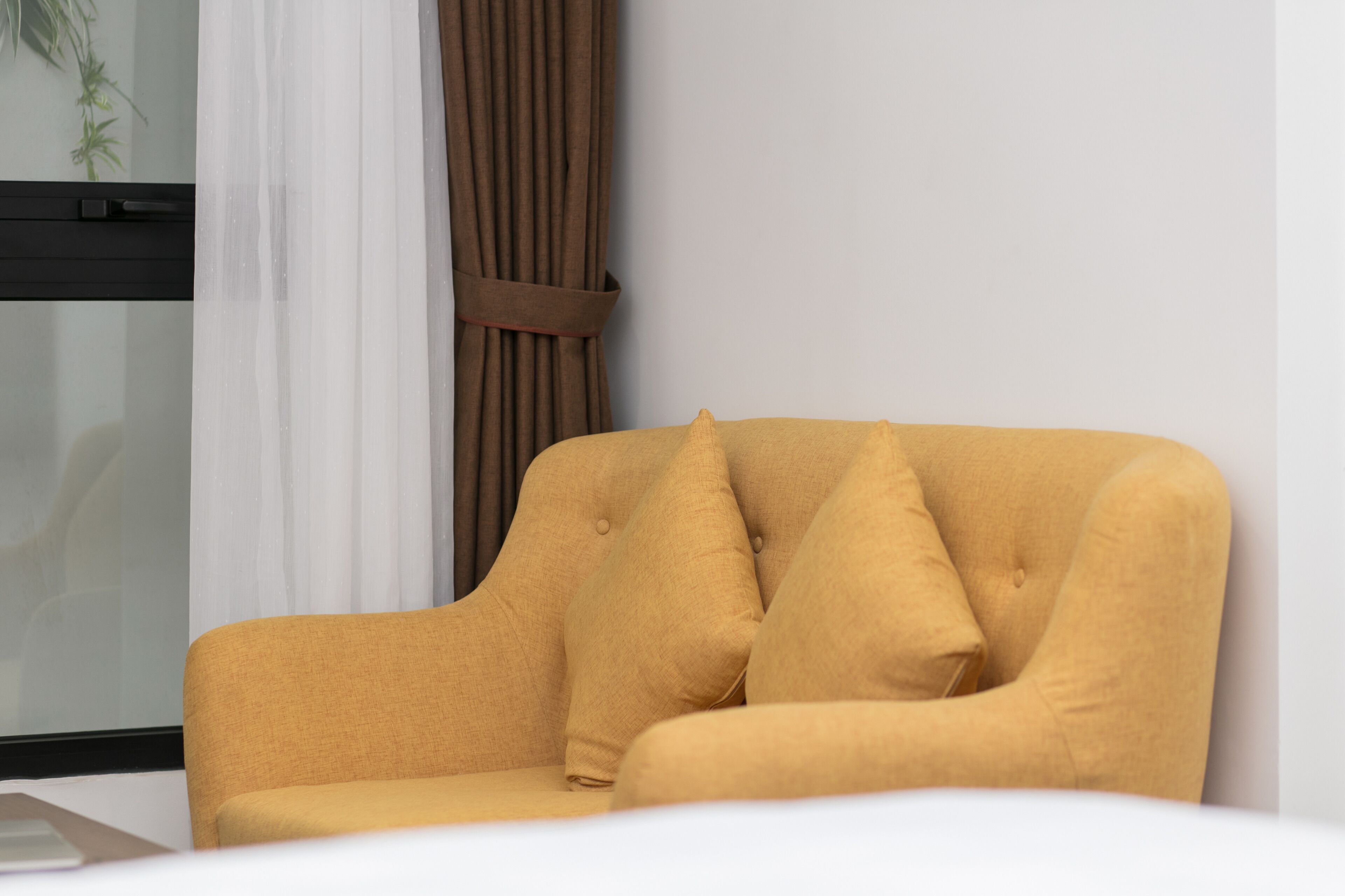 A mustard-colored sofa with a brown belt, positioned against a white wall with a window and curtains.