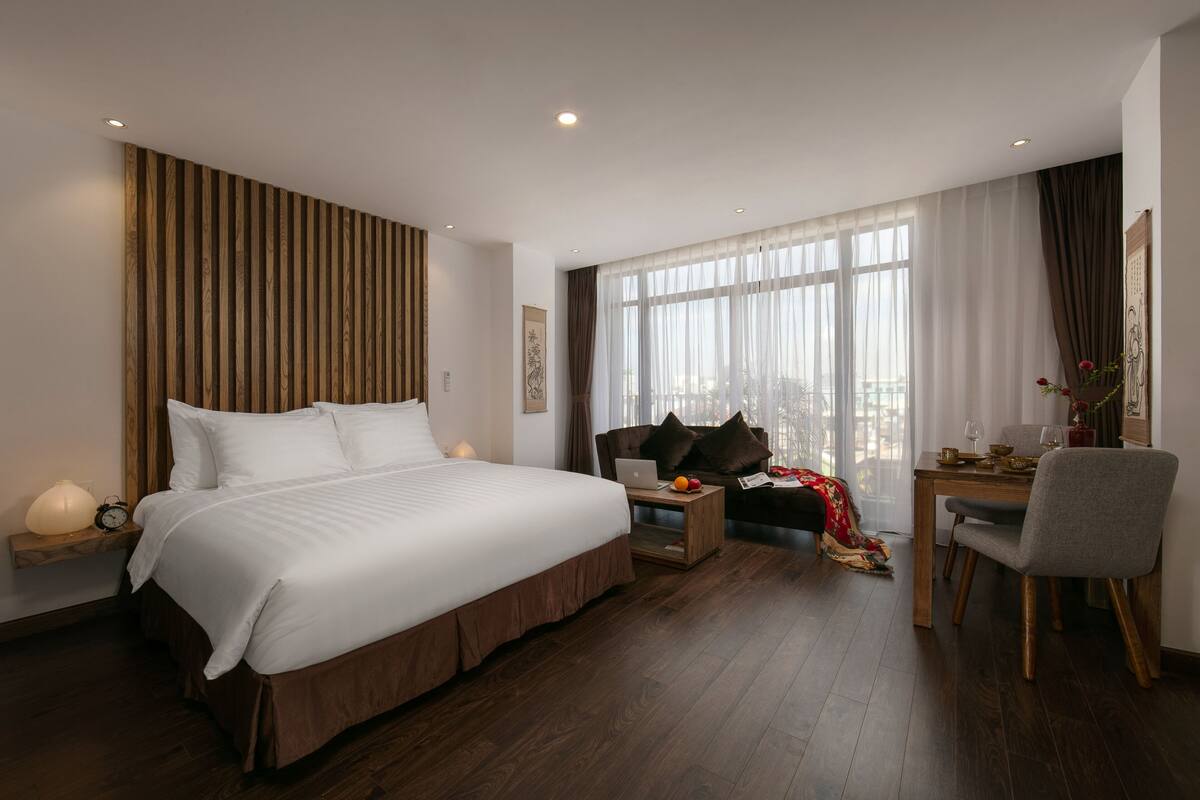 A hotel room with a large bed, a desk with a laptop, a chair, and a small table with a lamp.