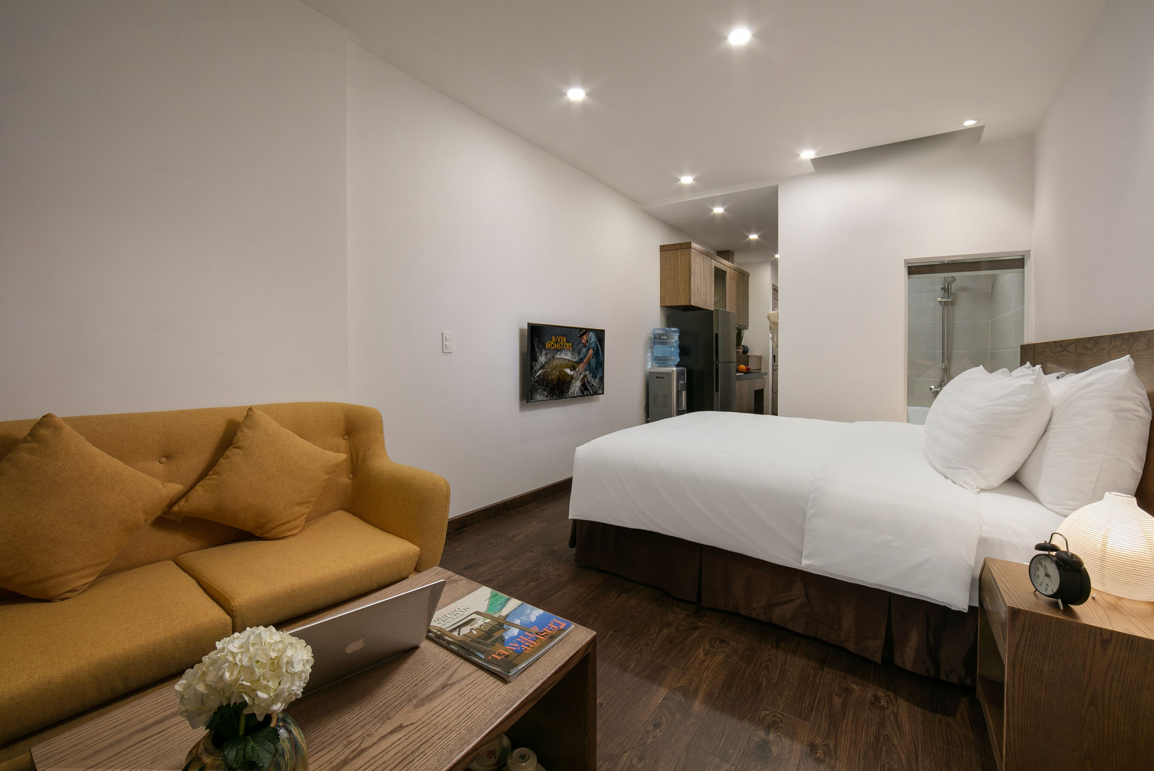 A hotel room with a bed, a sofa, a wooden coffee table, a laptop, and a water bottle.