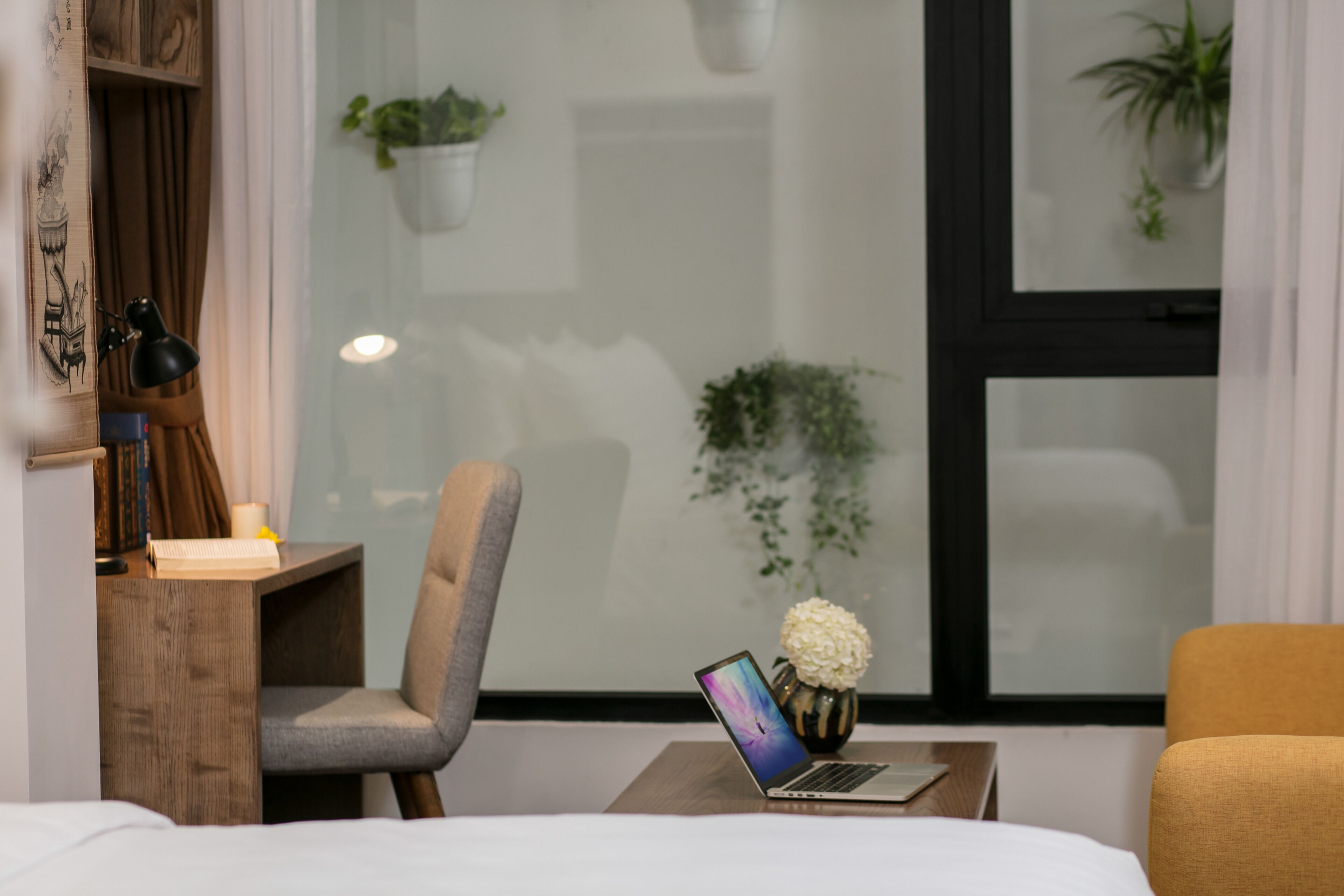 A modern hotel room with a bed, a desk with a laptop, a chair, and a small table with a vase of flowers.