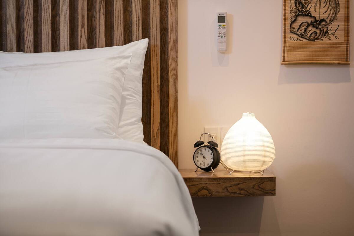 A neatly made bed with white linens, a bedside table with a black alarm clock and a lit paper lantern, and a wall-mounted thermostat.