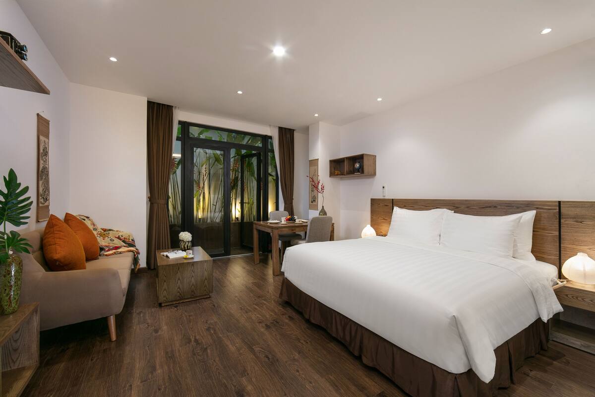 A modern hotel room with a large bed, a sofa, a small table, and a desk. There is a window with curtains and a view of greenery outside.
