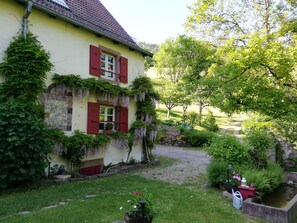 Une maison avec des volets rouges, un jardin avec un étang et un chemin menant à la maison.