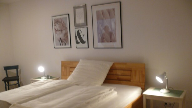 Een keurig opgemaakt bed met een houten hoofdbord, twee nachtkastjes met lampen, en drie ingekaderde foto's aan de muur.