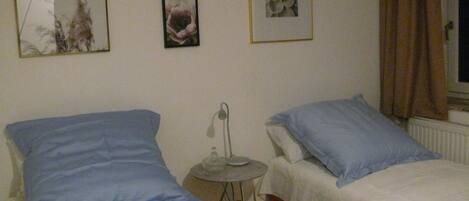 A room with two beds, each with blue and white bedding. There are three framed pictures on the wall, one of a dandelion, one of a flower, and one of a rose. A small table with a lamp is in the center of the room.