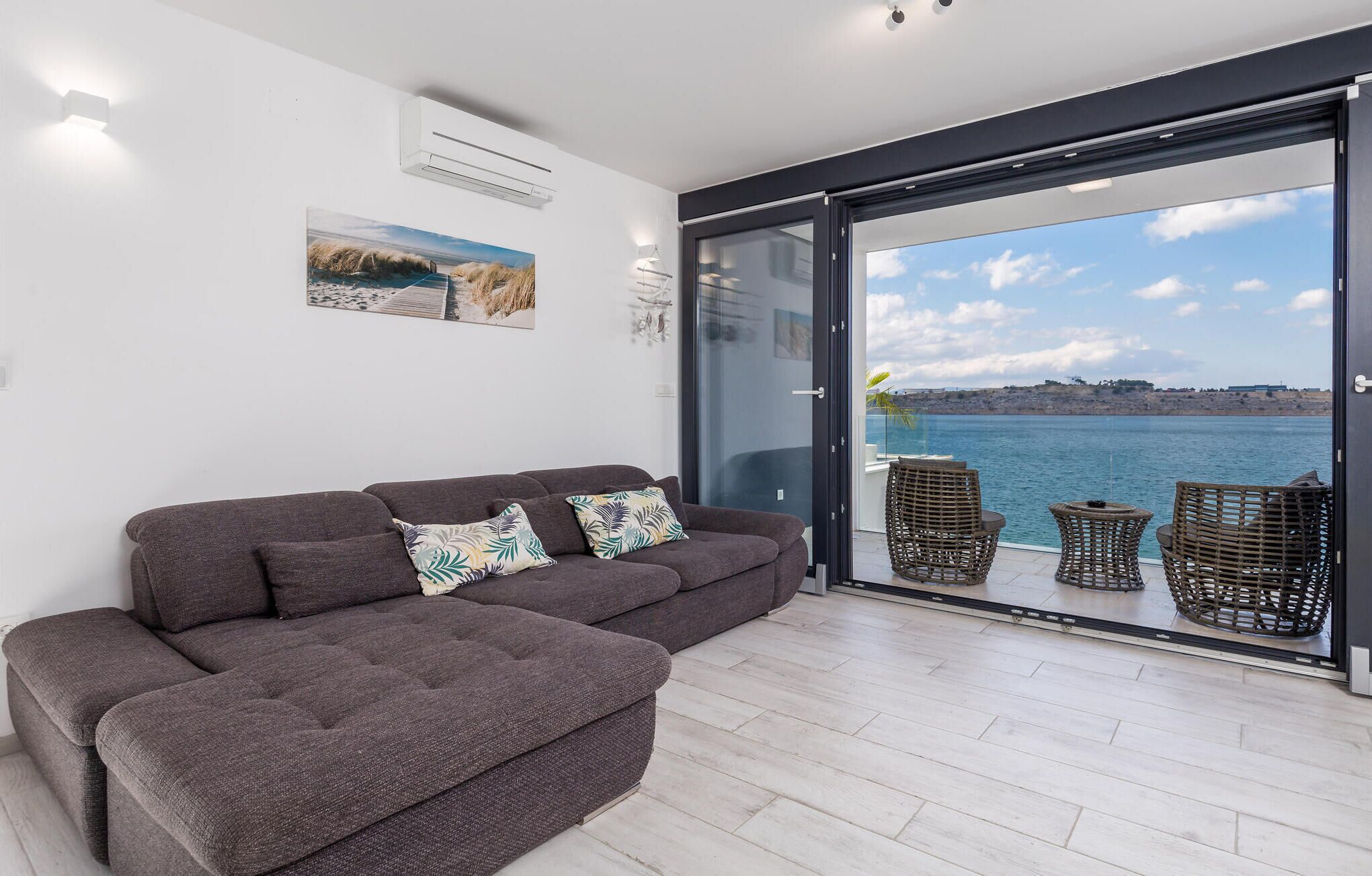 Un salon moderne avec un canapé d’angle gris, deux fauteuils en osier et une porte vitrée donnant sur un balcon avec vue sur la mer.