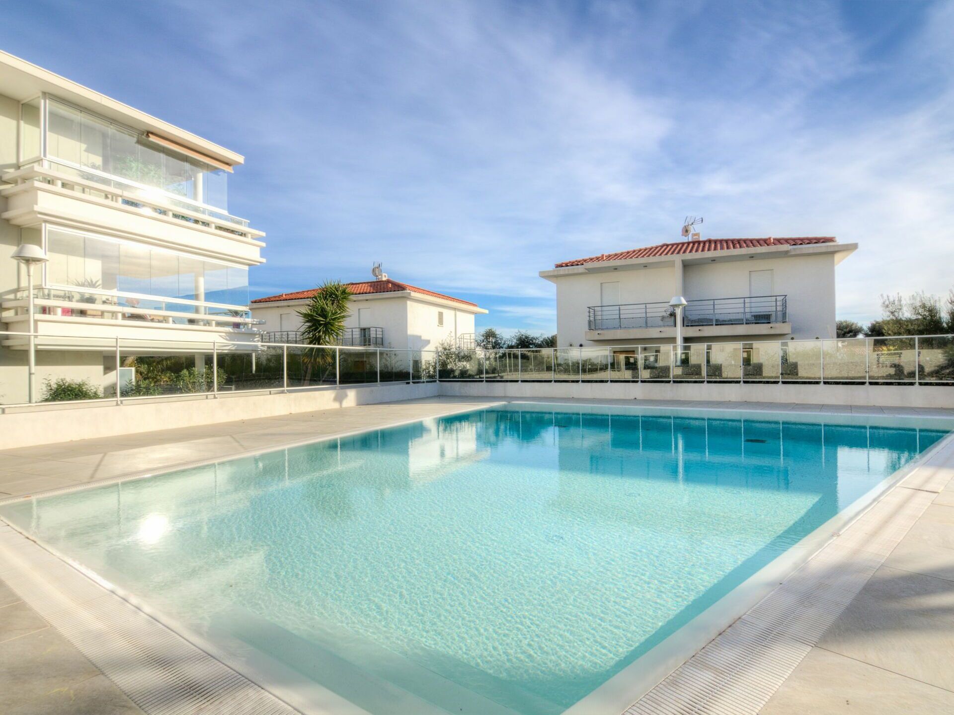 A swimming pool with clear blue water, surrounded by a modern building with balconies and a red-tiled roof.